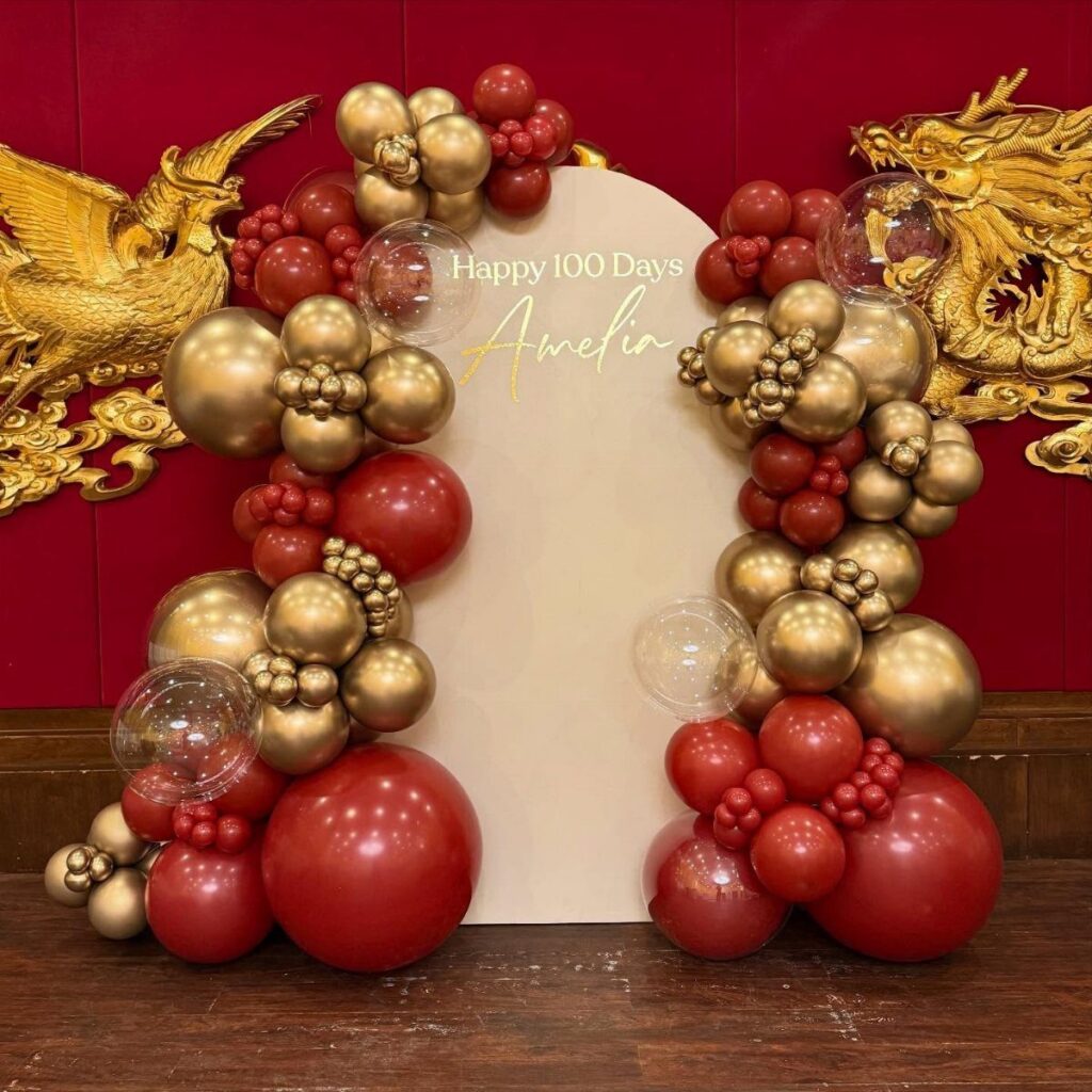 Balloon arch in red and gold surrounding a 100 days celebration backdrop