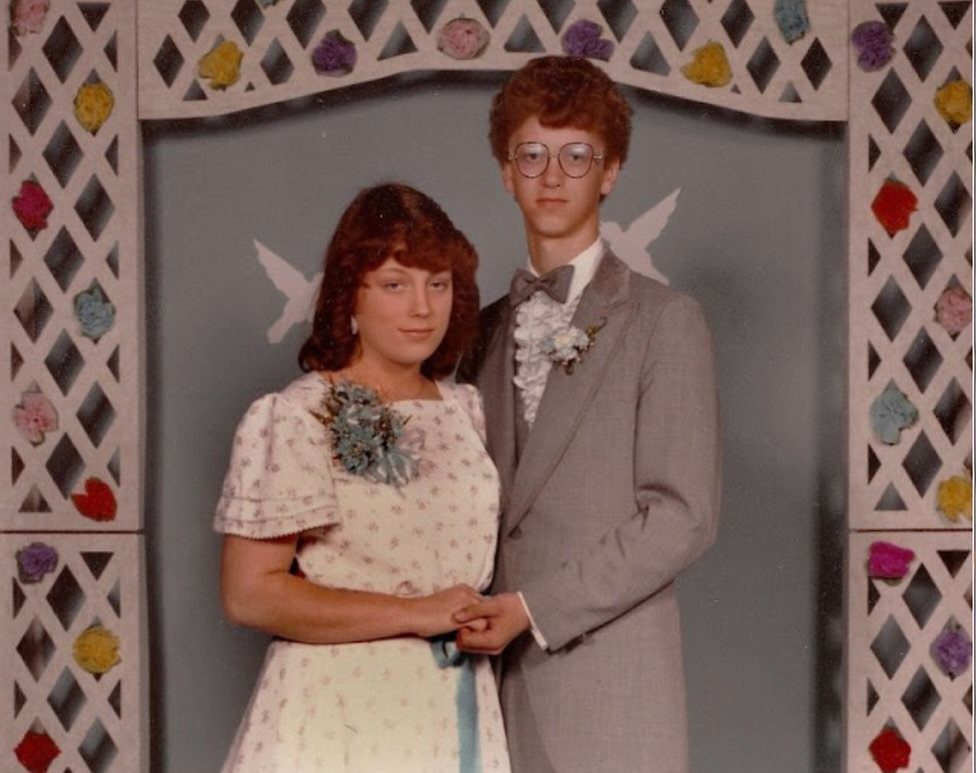 Young couple in 80s prom attire posing against a white lattice floral backdrop.
