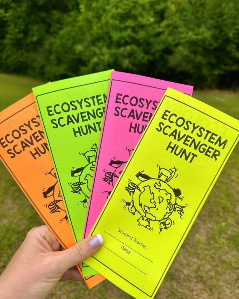 Neon colored ecosystem scavenger hunt worksheets held against a blurred green outdoor background.