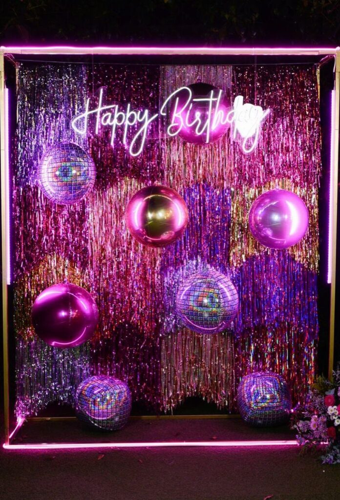 Disco party backdrop with purple tinsel, mirrored disco balls, and neon birthday sign.