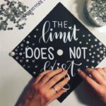 Hands decorating a black graduation cap with white lettering and small white pearls.