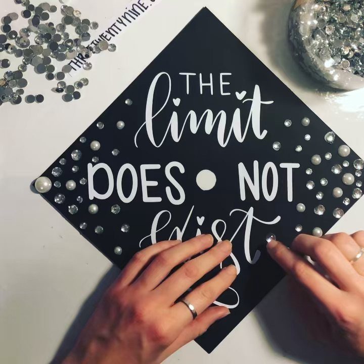 Hands decorating a black graduation cap with white lettering and small white pearls.