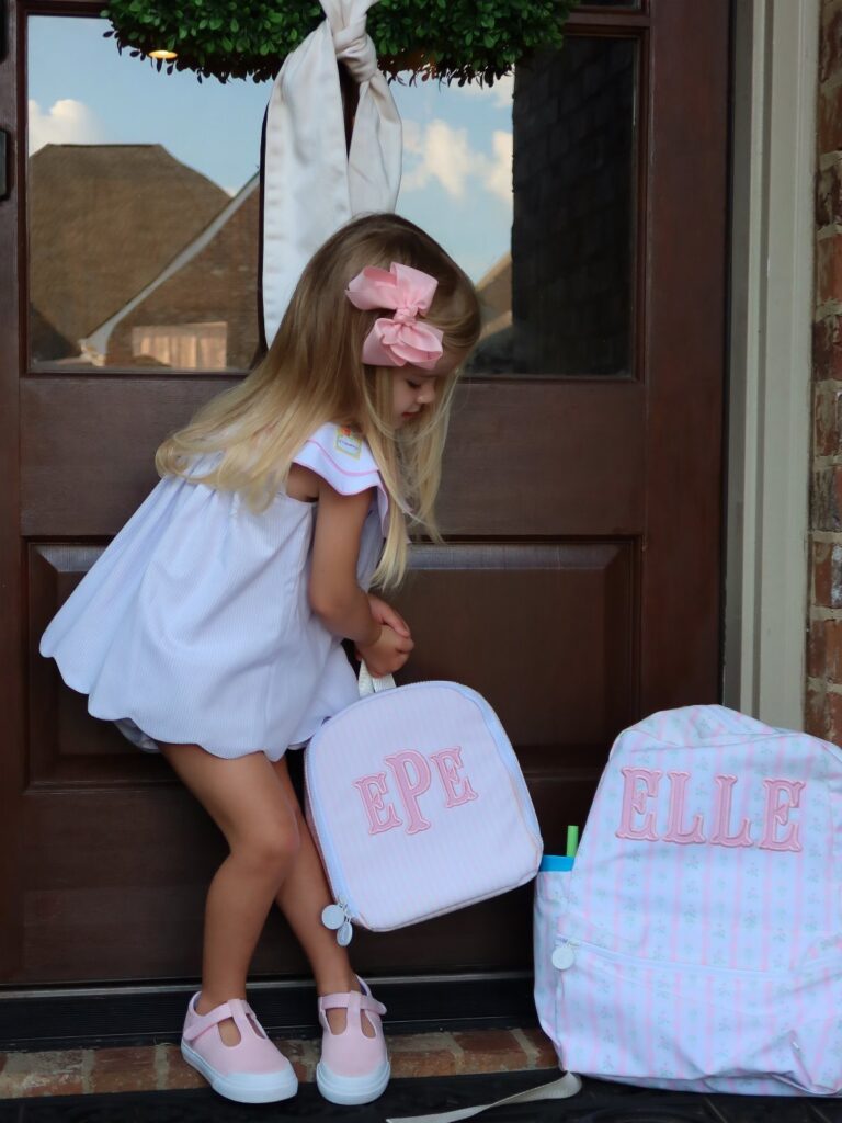 Young girl in a white dress and pink bow holding a personalized backpack.