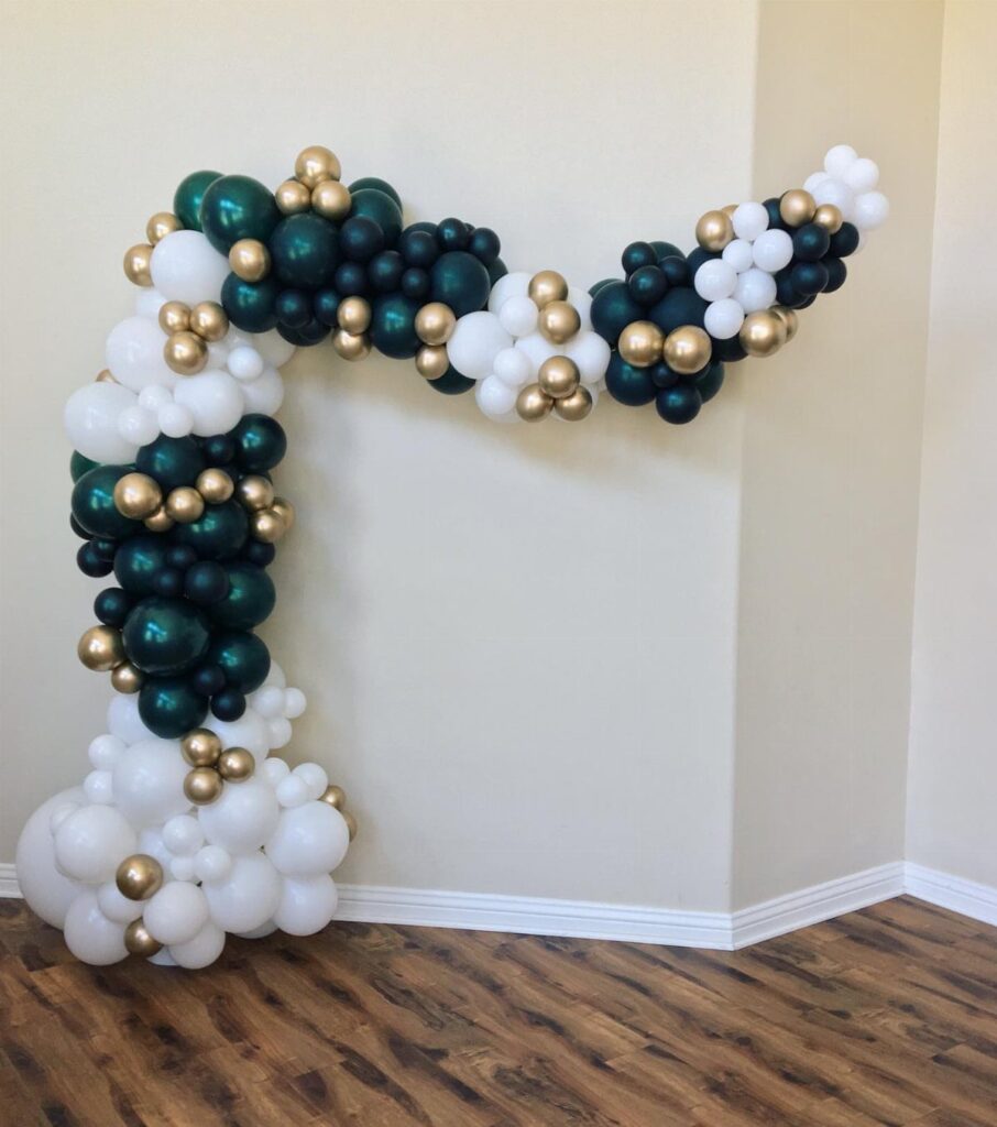 Dark green, white, and gold balloon arch curving against a beige wall.