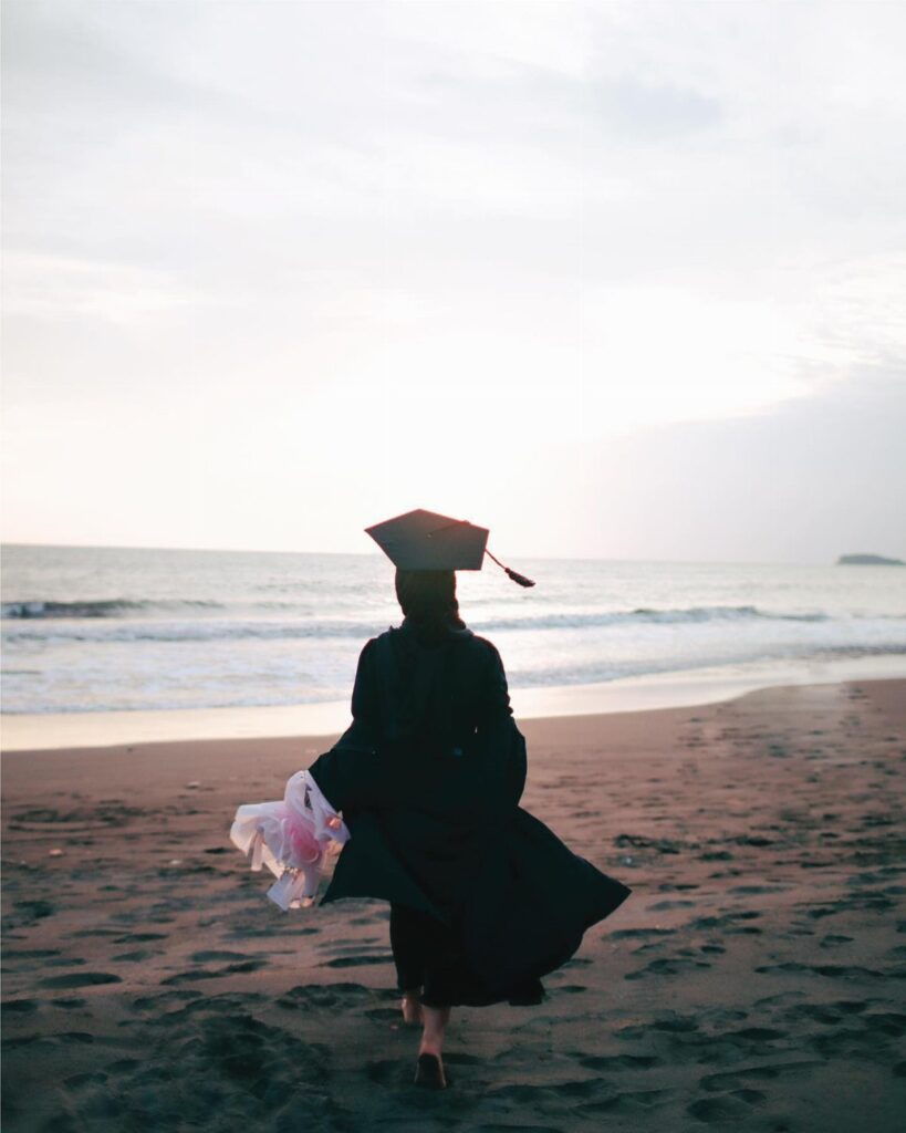 Graduate in cap and gown walking on a sandy beach toward the ocean.