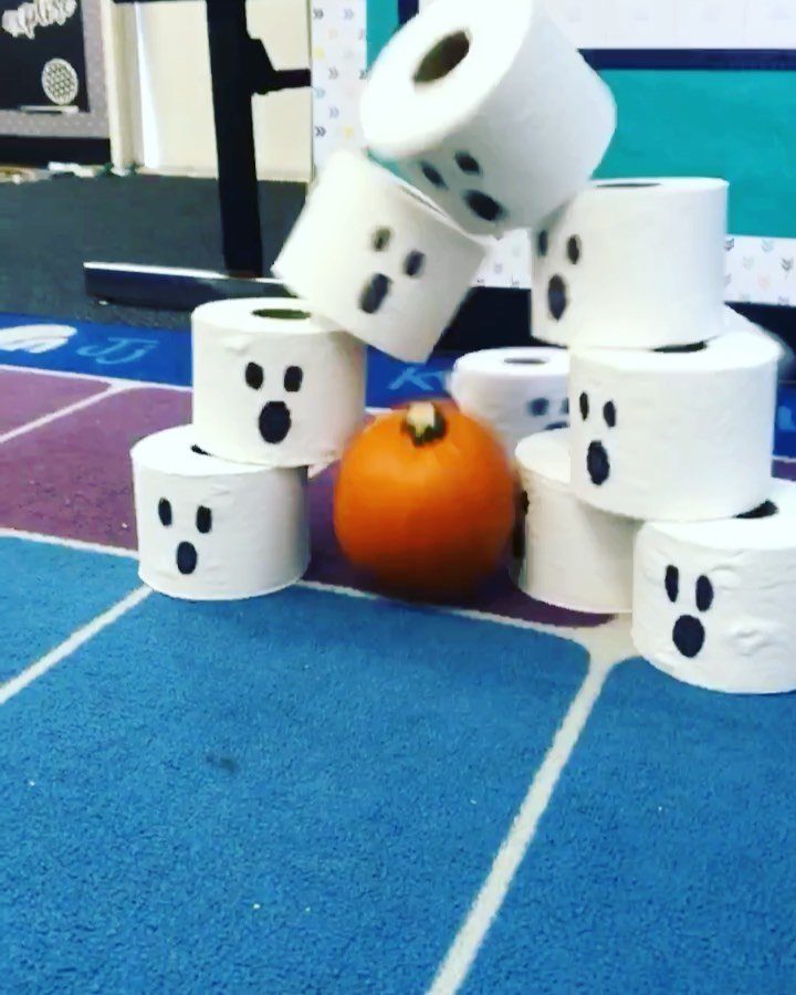 Mini pumpkin knocking over toilet paper rolls decorated as ghosts for bowling game.