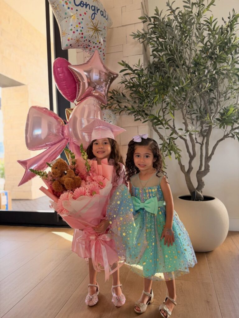 Two young girls celebrating, one wearing a pink graduation cap and holding flowers.