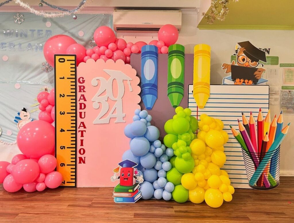 Graduation backdrop with colorful balloons, giant crayons, a ruler, and 2024 signage.