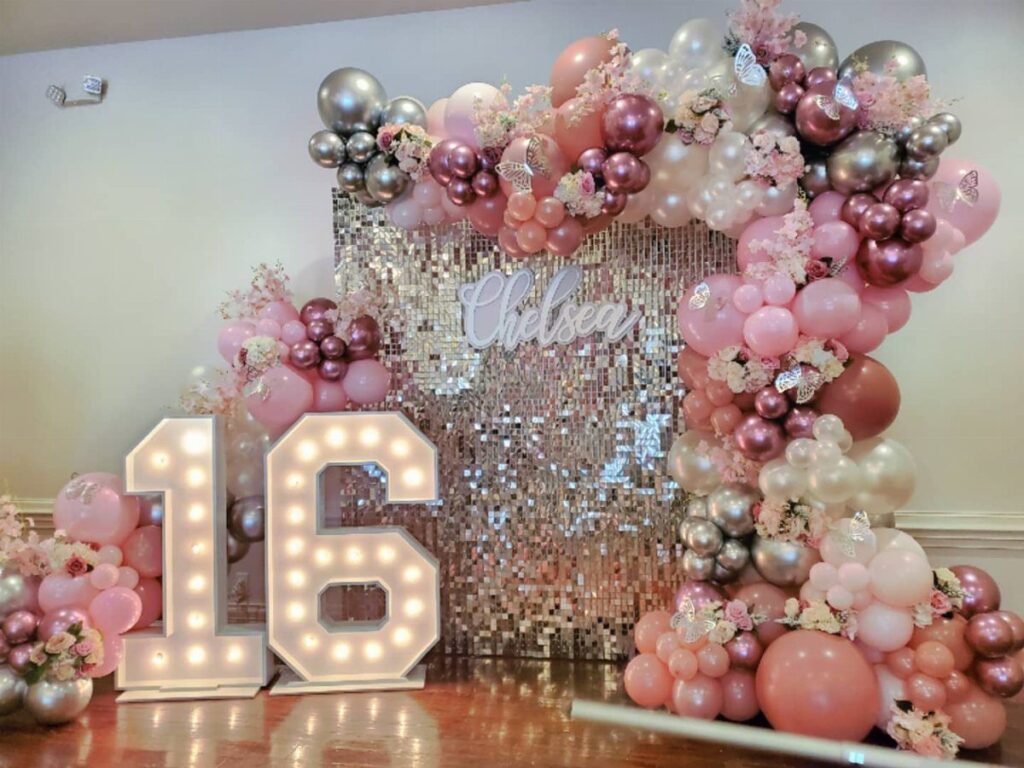 Illuminated number 16 marquee signs beside a pink and silver balloon arch backdrop.