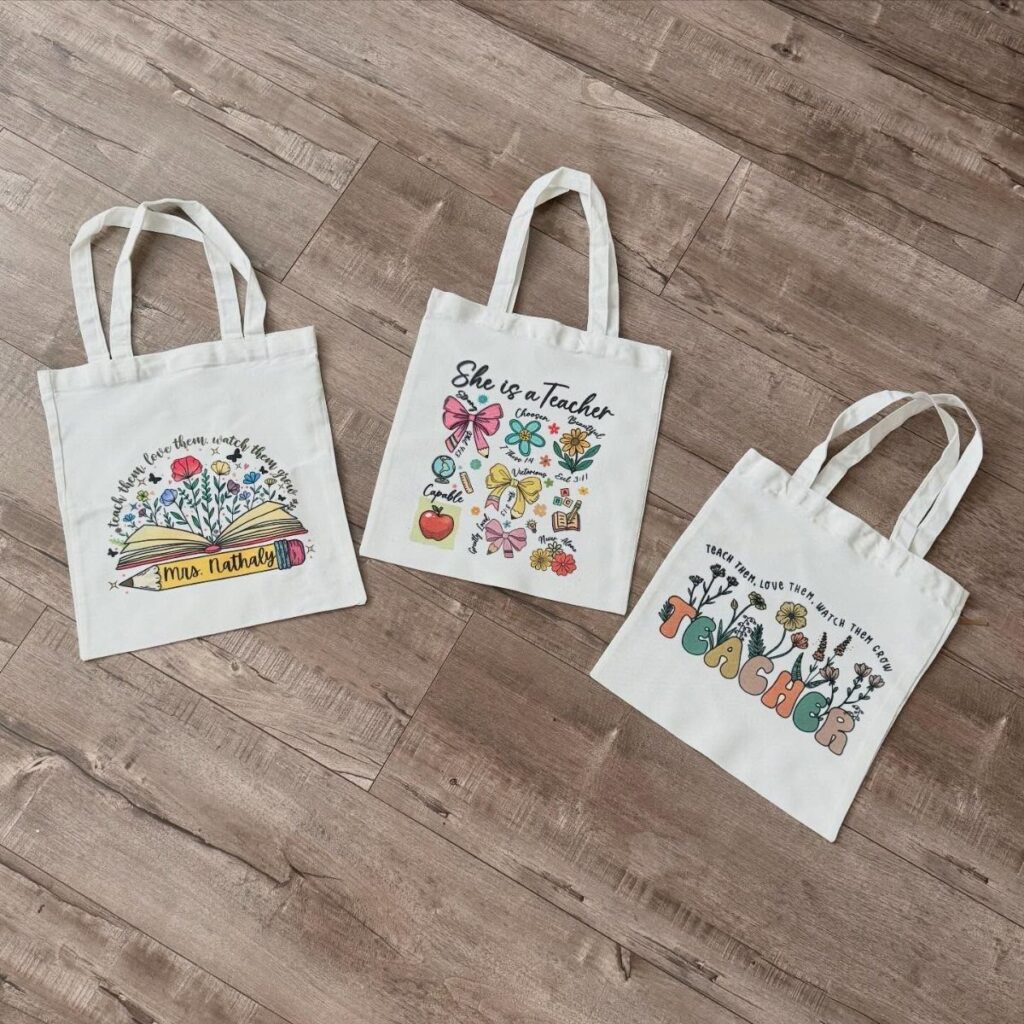Three white canvas tote bags with teacher-themed illustrations on a wooden floor.