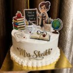 White frosted cake decorated with teacher-themed toppers and the name Mrs Sheikh.
